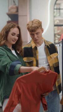 Vertical Screen. Clothing Store Young People Shopping. Happy Couple Browsing Racks Together, Discussing Choices and Holding up a Colorful Garment While Smiling, Enjoying a Weekend Retail Shopping Date