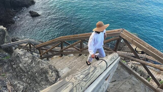 Woman dressed in white with wide brimmed hat walks down the wooden staircase along a coastal hiking trail in Riomaggiore, Cinque Terre in Italy