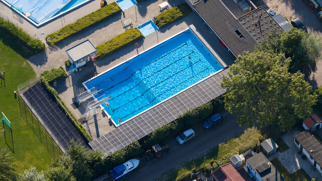 Aerial view of an outdoor swimming pool with swimming lanes and diving boards, surrounded by paved areas, trees, and parked cars in Lauwerzijl, Groningen, Netherlands.