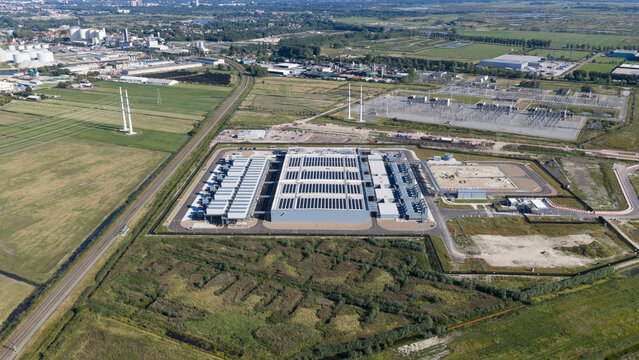 Aerial view of a large data center facility with solar panels on the roof, surrounded by green fields, power infrastructure, and railway tracks in Groningen, Groningen, Netherlands.