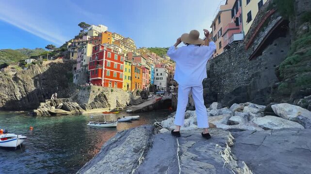 Tourist woman with hat posing at colorful Riomaggiore pier during summer vacation in the Cinque Terre, Italy