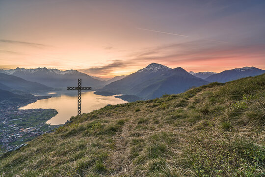 Aerial view of a large metal cross standing on a grassy hillside overlooking the calm waters of Lake Como at sunset in Musso, Lombardy, Italy.