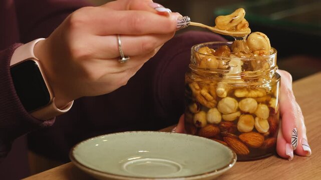 Cafe worker scoops honey nuts from glass jar on wooden cafe counter. Woman lifts glossy topping with spoon preparing sweet garnish slowly