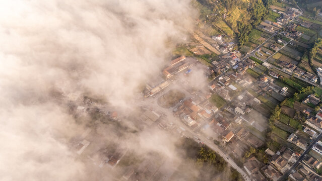 Aerial view of Selo village partially obscured by morning clouds with terraced fields and residential rooftops visible below, Selo, Central Java, Indonesia.