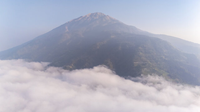 Aerial view of the volcanic peak of Mount Merbabu rising above a thick blanket of clouds with lush green slopes in Selo, Central Java, Indonesia.