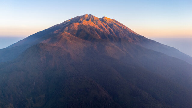 Aerial view of Mount Merbabu at sunrise with golden light illuminating the peak and slopes, surrounded by lush vegetation in Selo, Central Java, Indonesia.