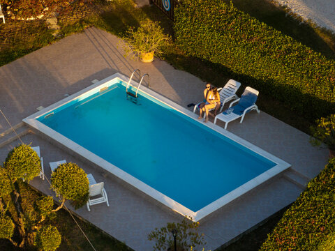 Aerial view of a rectangular swimming pool with two people relaxing on lounge chairs on a tiled patio surrounded by green hedges in Montelparo, Marche, Italy.