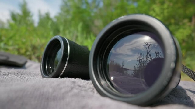 Field binoculars rest on a soft blanket during a sunny day, mirroring the open sky and reed grasses in the glass element. The optical device offers precise focus for outdoor surveillance.