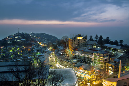 Aerial view of Murree at dusk, featuring illuminated buildings, snow-covered rooftops, and a mosque minaret nestled in the hilly landscape of Murree, Punjab, Pakistan.