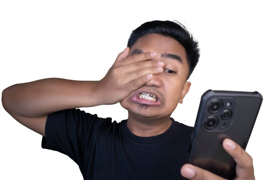 Frustrated and embarrassed Indonesian man wearing a black t-shirt looking at his mobile phone while covering his face with his hand in disbelief and cringing against a white background.