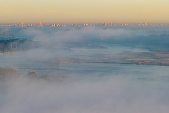 Aerial view of the Vistula river partially obscured by thick morning fog with a distant city skyline visible on the horizon under soft golden light in Poland.