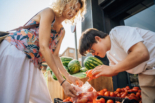 Mature mother and teenage son with Down syndrome choosing fresh tomatoes at outdoor market in Spain