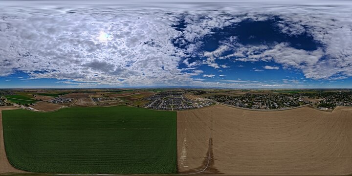 Aerial view of agricultural fields and suburban residential neighborhoods under a cloudy blue sky in Johnstown, Colorado, United States.