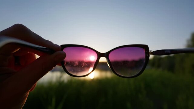 Hand holds stylish sunglasses against a setting sun over a calm lake with grassy shoreline. The tinted violet lenses offer an evocative visual framing device for summer travel stories.