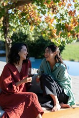 Fototapeta premium Two women laughing with glasses of wine by the pool under autumn vines for warm social background and lifestyle banner design