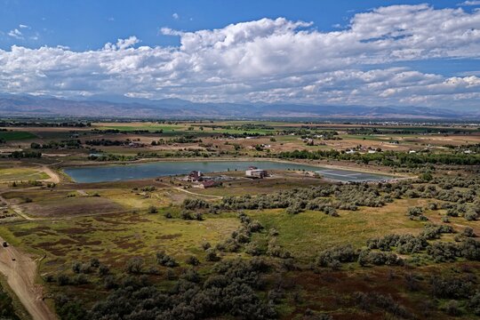 Aerial view of a reservoir surrounded by open fields, shrubs, and distant mountains under a blue sky with white clouds in Erie, Colorado, United States.