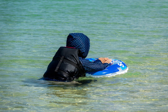 Woman in a black life jacket and polka-dot hood floating on a blue inflatable ring in the shallows of Hoshizuna beach, Yaeyama Islands, Iriomote, Japan