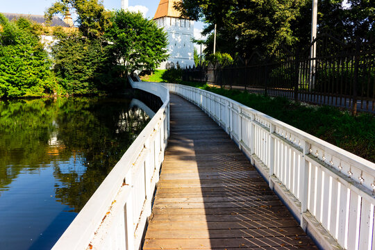 Scenic wooden walkway stretching along the tranquil pond leading towards the white stone towers of the historic Pafnutiev Borovsky Monastery on a sunny summer day in Russia