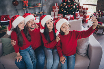 Naklejka premium Family taking Christmas selfie on couch wearing Santa hats and matching red sweaters with decorated tree and wrapped gifts in cozy living room