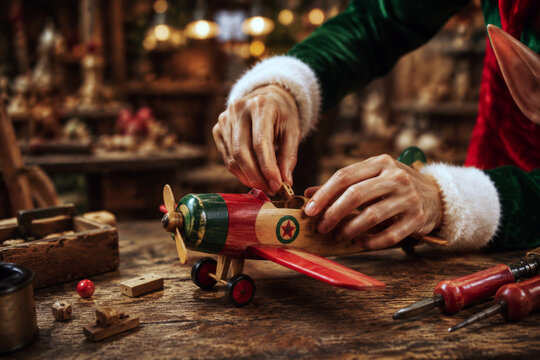 Close-up hands assemble a small wooden airplane with hand tools on a workshop bench surrounded by warm festive lights. Detailed toy craftsmanship. New Year and Christmas concept