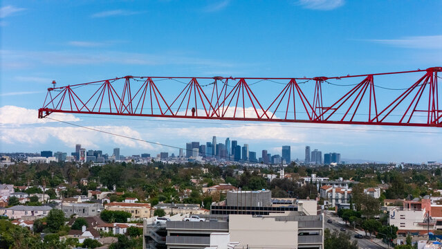 Aerial view of a red construction crane jib extending over the Los Angeles skyline with residential buildings and trees in the foreground in Los Angeles, California, United States.