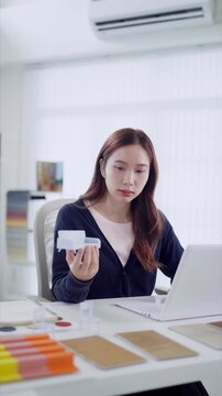 Young Asian designer examines miniature furniture model while working on laptop in bright studio