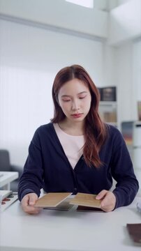 Young Asian interior designer comparing wood veneer swatches at desk in bright modern studio
