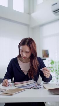 Young Asian interior designer examines fabric swatches and miniature chair model at bright studio desk