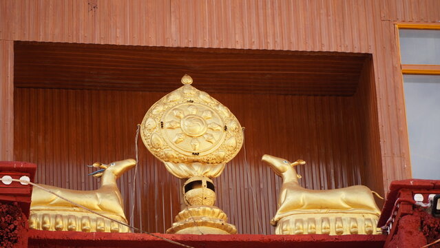 Golden deer statues flanking a decorative wheel above a red wooden structure with golden accents in bright sunlight on a clear day in Ladakh, India