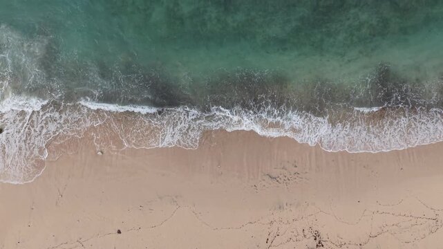 High-angle aerial photo of a wide sandy beach with foamy ocean waves rolling onto the shoreline, scattered footprints and seaweed, conveying calm, travel and natural coastal beauty