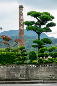 Brick chimney painted with vertical Japanese lettering rising above manicured pine trees and a distant mountain backdrop, Kyushu region, Arita, Japan