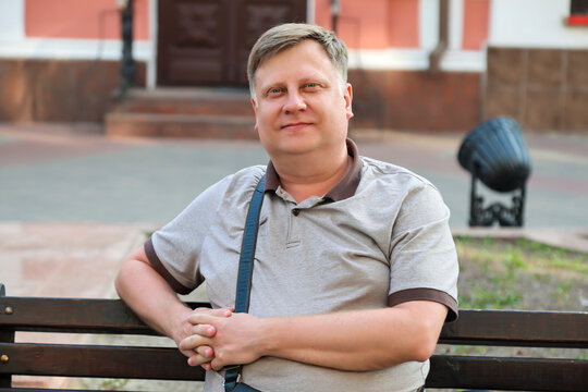 A mature blond man sits happily on a park bench, enjoying rest and fresh air during the day.