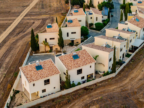 Aerial view of modern residential houses with tiled roofs and solar water heaters surrounded by dry landscape in Alethriko, Larnaca, Cyprus.