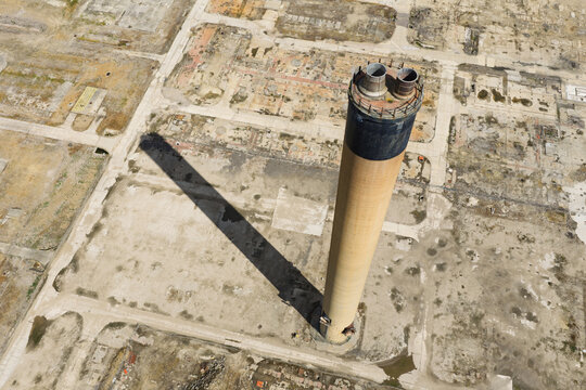 Aerial view of a tall industrial chimney casting a long shadow over a cleared and desolate industrial site in the United Kingdom.