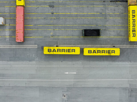 Aerial view of yellow shipping containers labeled Barrier arranged on an asphalt surface with road markings and a red container, United Kingdom.