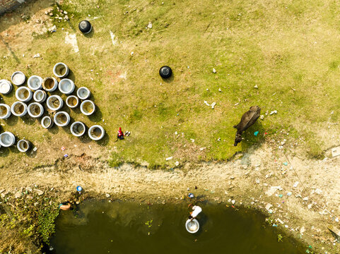 Aerial view of a water buffalo grazing on grass near a pond where people are washing pots, with numerous metal containers scattered on the ground in Dhaka, Dhaka Division, Bangladesh.