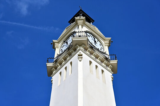 white stucco clock tower. the Port Authority of Valencia, Spain. beach area. large round white clocks. black handles. lush green palm tree. narrow vertical window. blue sky. travel , tourism concept 