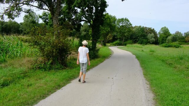 Young brunette woman walks along gravel road past lush green bushes and blooming summer wildflowers. Peaceful rural landscape of Croatia offers relaxation and slow living vibes.