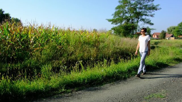 Young brunette woman walks along gravel road past lush green bushes and blooming summer wildflowers. Peaceful rural landscape of Croatia offers relaxation and slow living vibes.