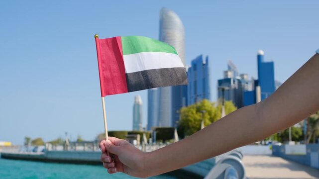 Female hand holding small UAE flag against Abu Dhabi skyline. Symbol of national pride and modernity with iconic skyscrapers in the background showcasing urban elegance and progress