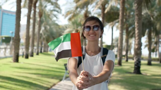 Woman tourist holding small UAE flag against Corniche Walking Trail in Abu Dhabi. Blend of nature, patriotism, and serenity, with lush greenery , views symbolizing harmony and progress.