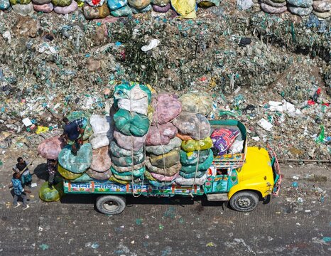Aerial view of a brightly colored truck heavily loaded with large sacks of waste, with workers loading more bags near a massive landfill site in Dhaka, Dhaka Division, Bangladesh.