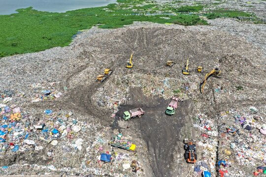 Aerial view of heavy machinery including excavators and dump trucks working at a large landfill site with piles of waste and surrounding green vegetation in Dhaka, Dhaka Division, Bangladesh.