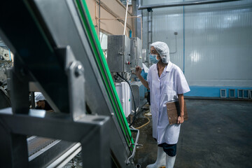 Fototapeta premium A food plant worker wearing a protective hairnet and face mask operates machinery control panels inside a large manufacturing factory with metal equipment and concrete floors.