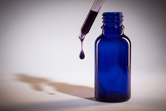 A purple liquid drop falls from a glass pipette into a small empty blue bottle with against neutral background pipette positioned above hangs glass