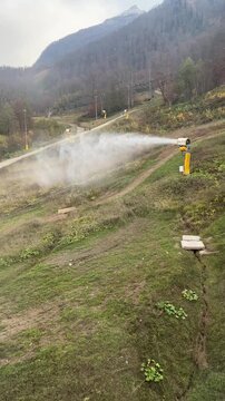 A snow-making machine operates on a ski slope. The machine sprays water vapor into the air, creating artificial snow. The background features a mountainous landscape.