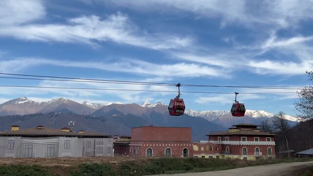 A scenic view of two red gondolas traveling over a mountainous landscape. Snow-capped peaks and a clear blue sky are visible in the background.