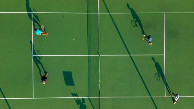 Aerial view of four people playing a tennis match on a green court with long shadows cast by the players in Kecamatan Prambanan, Jawa Tengah, Indonesia.
