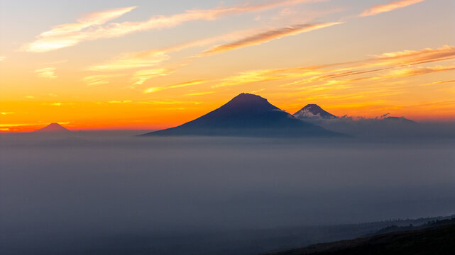 Aerial view of Mount Sumbing volcano rising above a thick layer of clouds during a vibrant golden hour sunset in Cangkringan, Yogyakarta, Indonesia.