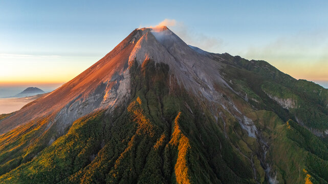 Aerial view of Mount Merapi with volcanic smoke rising from the peak, illuminated by sunrise light over lush green slopes in Cangkringan, Special Region of Yogyakarta, Indonesia.